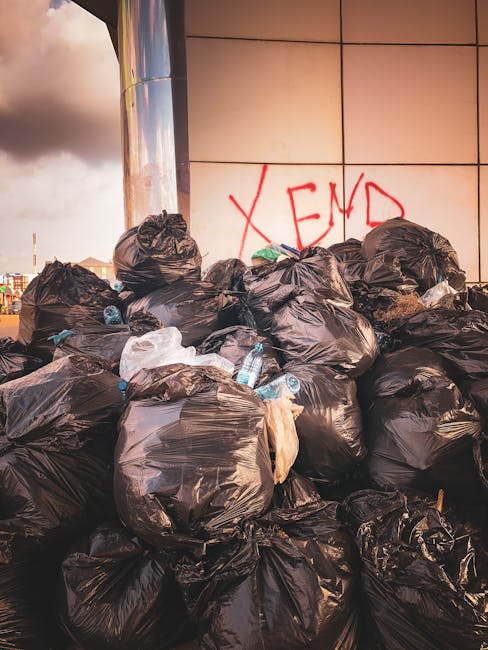 A large pile of black rubbish bags, some featuring transparent areas revealing white waste or paper inside, are stacked against a building with a metallic and tiled wall surface. The bags appear fille