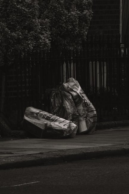 A large pile of black rubbish bags, some featuring transparent areas revealing white waste or paper inside, are stacked against a building with a metallic and tiled wall surface. The bags appear fille