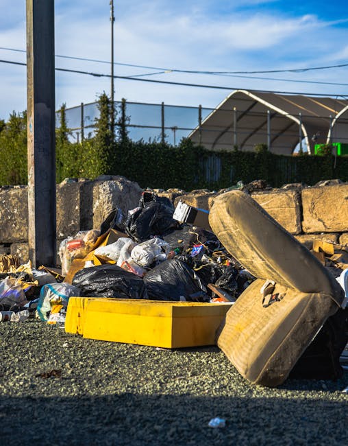A pile of mixed waste materials situated on a paved outdoor surface. The waste includes black plastic garbage bags, some of which are torn open, revealing discarded packaging and trash inside. There i