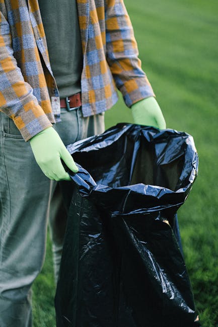 A pile of mixed waste materials situated on a paved outdoor surface. The waste includes black plastic garbage bags, some of which are torn open, revealing discarded packaging and trash inside. There i